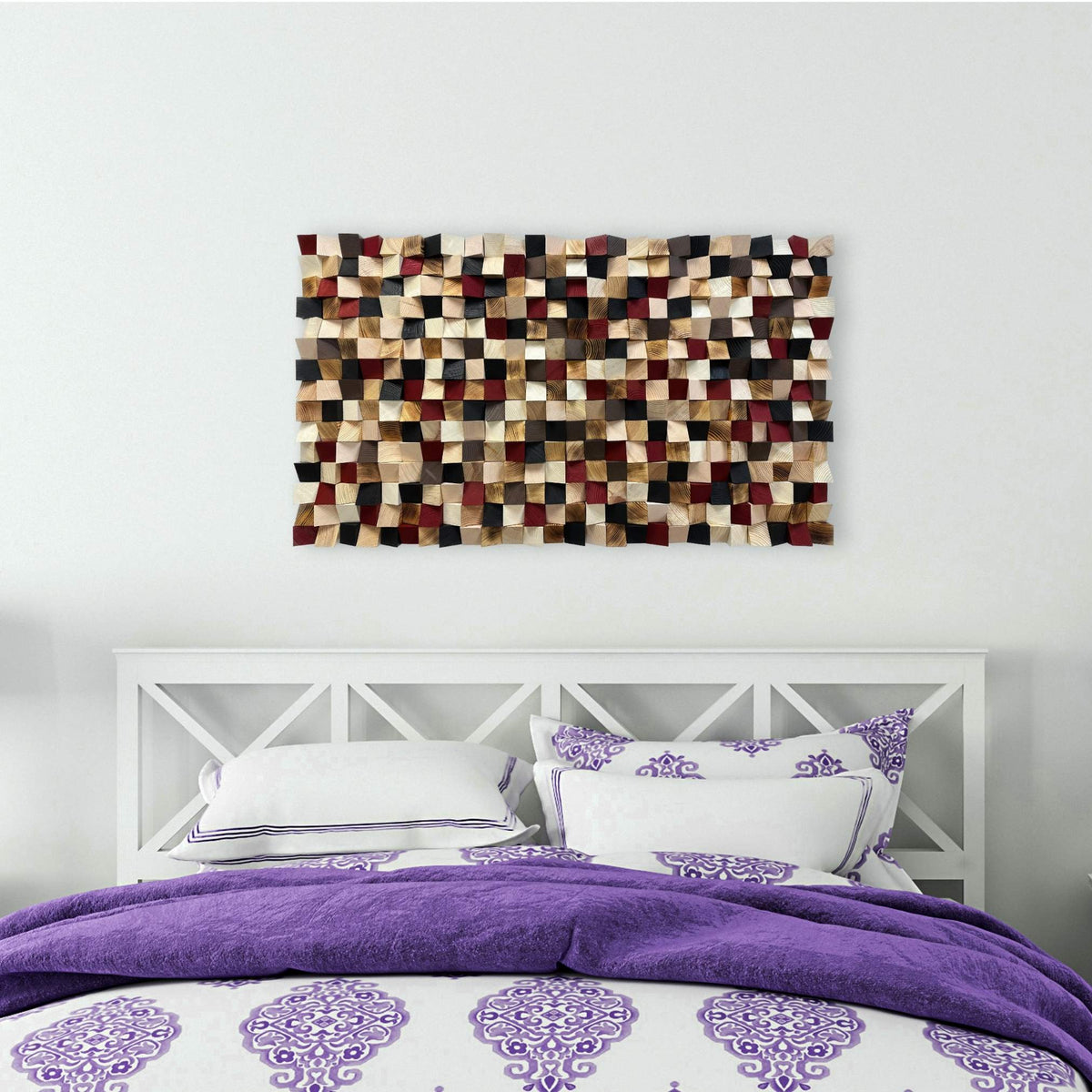 A modern wall art piece featuring a textured mosaic of rectangular patches in beige, brown, red, and black, mounted on a white wall above a bed with purple and white patterned bedding.