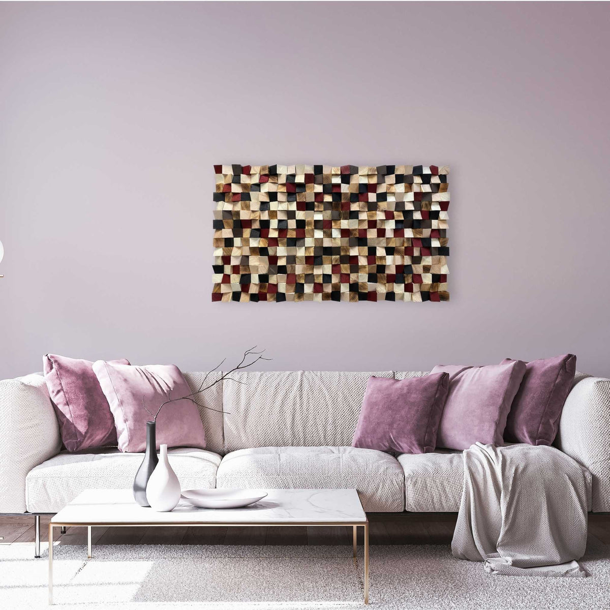 A modern living room setting featuring a rectangular woven wall art piece with a multicolor geometric patchwork pattern in beige, brown, black, and red, hanging on a lavender-gray wall above a light gray sofa with purple pillows.