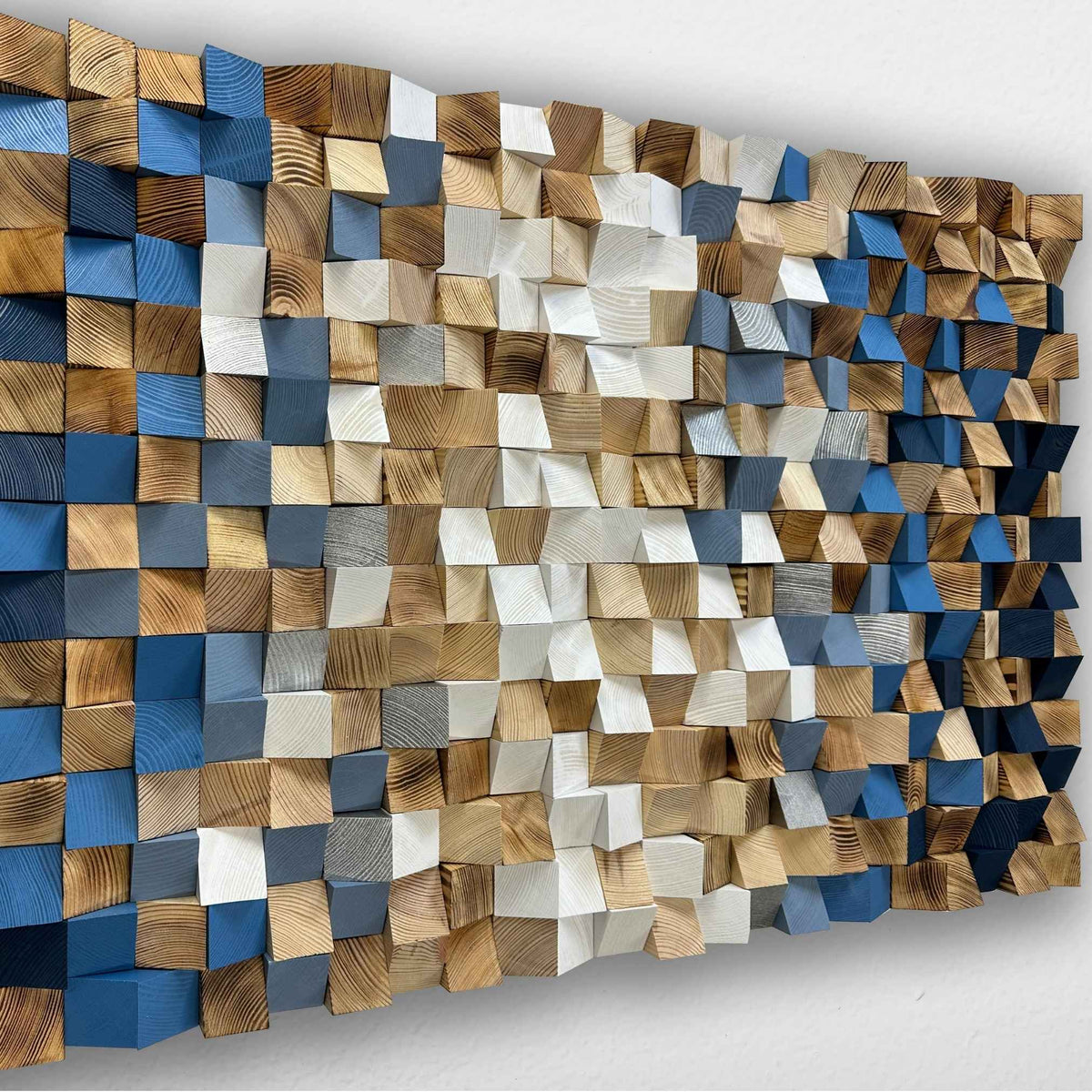 A rectangular wooden wall art piece made of many small cuboid blocks in mixed natural wood tones, blue, gray, and white, mounted against a plain light wall.