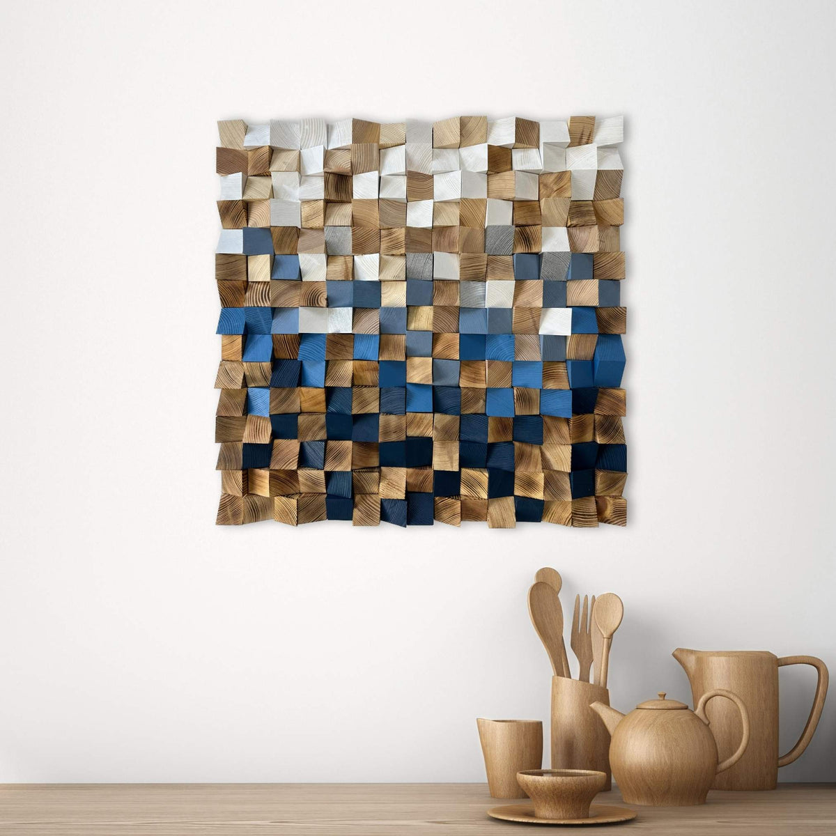 A square wall-mounted wooden mosaic art piece with a pixelated block pattern in shades of brown, beige, gray, and blue, hanging on a white wall above a wooden table with ceramic kitchenware.