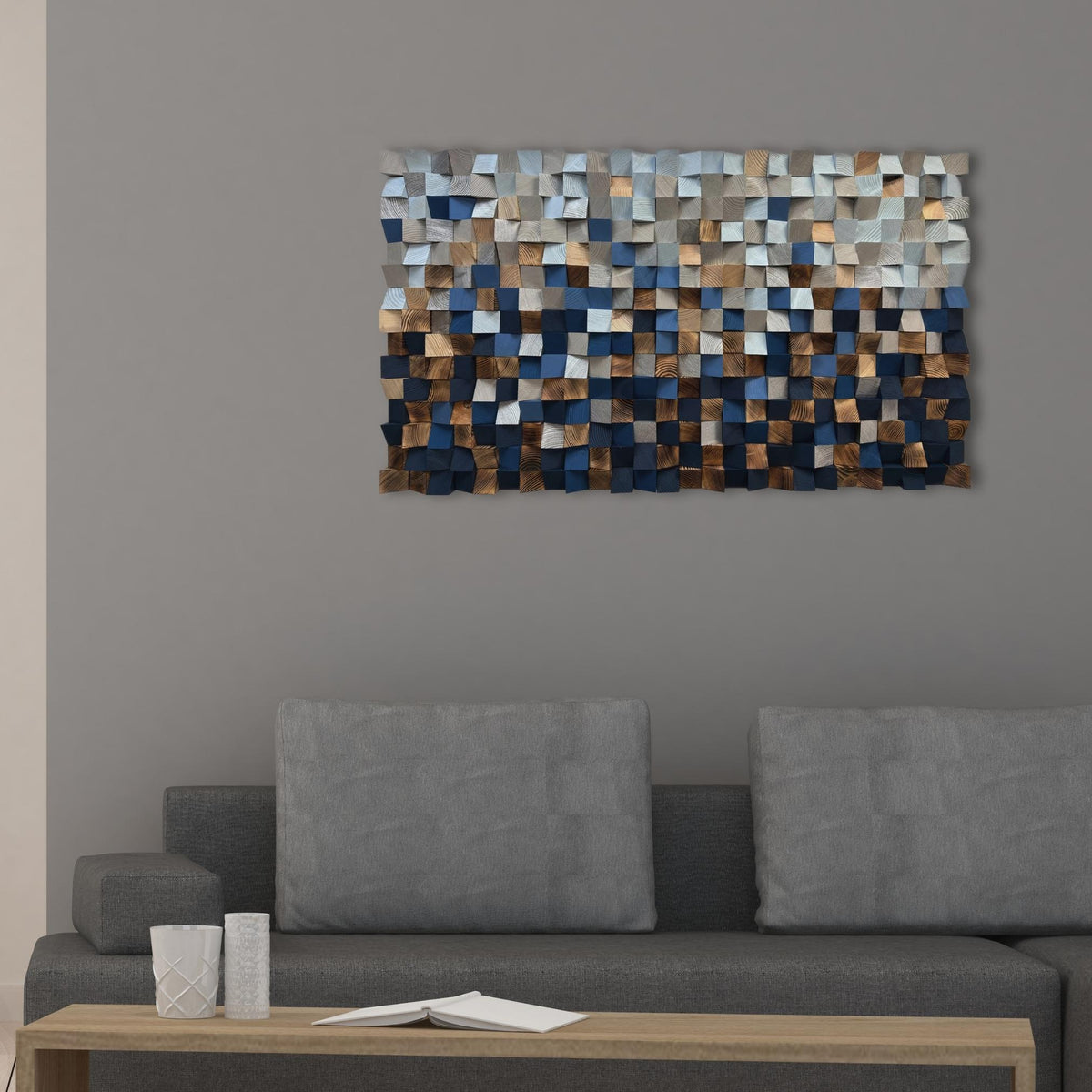 A rectangular wall art piece with a mosaic of textured square tiles in shades of blue, gray, beige, and brown, mounted on a gray wall above a modern gray sofa.