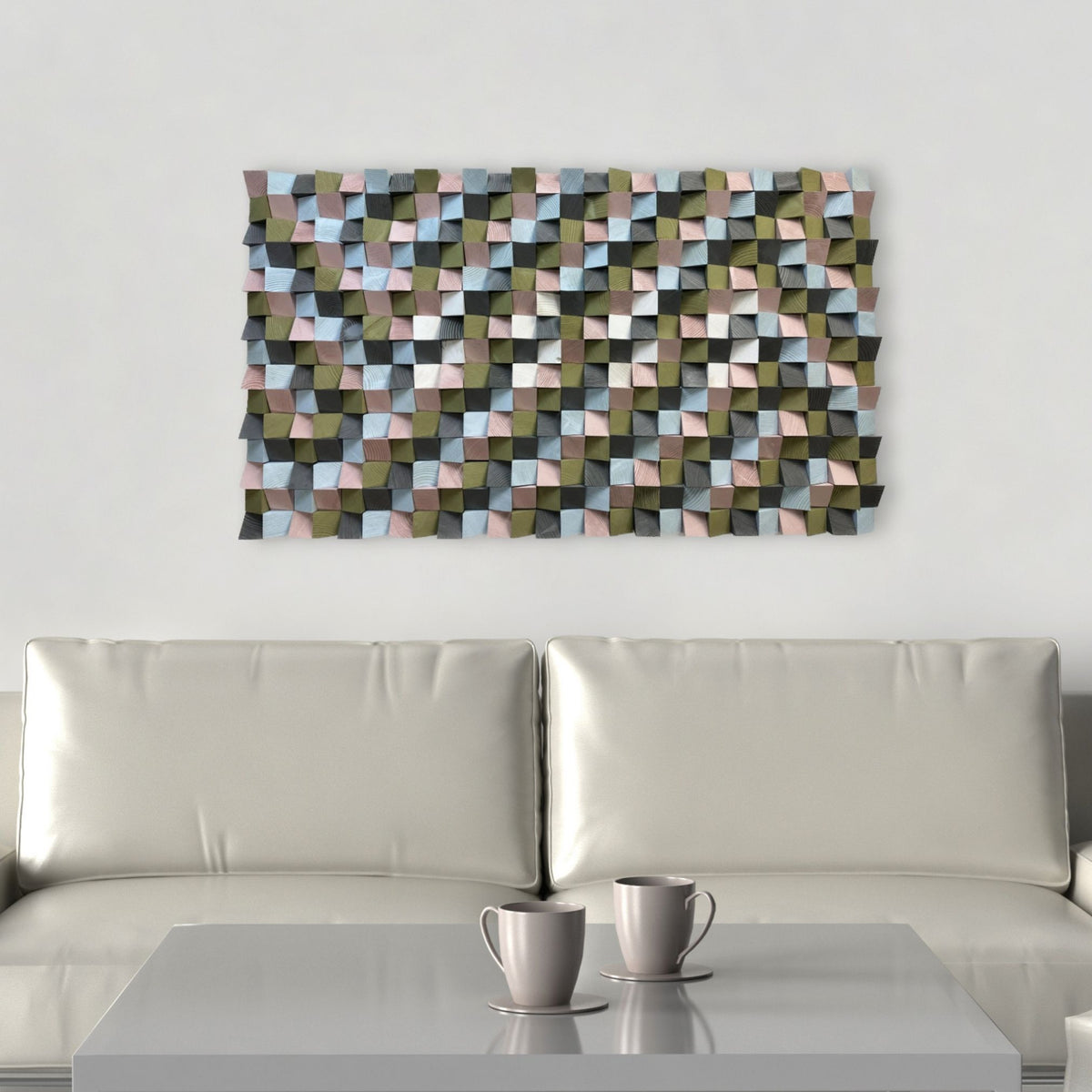 A rectangular wall art piece with a 3D geometric cube pattern in muted tones of olive green, dusty pink, light blue, and charcoal, displayed above a modern white leather sofa with two gray cups on a table in the foreground.